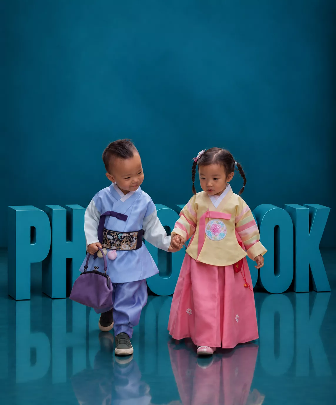 청록색 PHOTOBOOK 조형물 앞에서 한복을 입고 손잡고 걷는 두 아이의 성장앨범 홍보 이미지