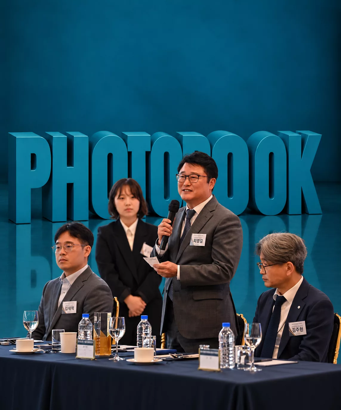청록색 PHOTOBOOK 조형물 앞에서 마이크로 발언하는 남성과 테이블 주변 참석자들의 기업행사앨범 홍보 이미지